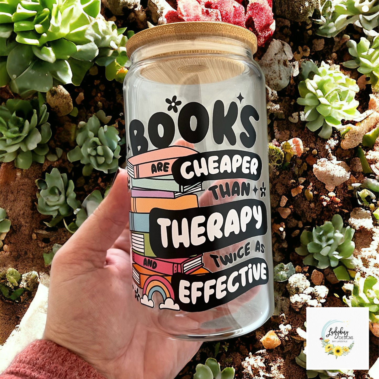 Book are Cheaper than Therapy Glass Can 16oz with Bamboo Lid and Plastic Straw. Ideal for iced coffee and perfect as a book lover&#39;s gift. Shown on a cozy reading table with a book and coffee cup, highlighting its stylish design.