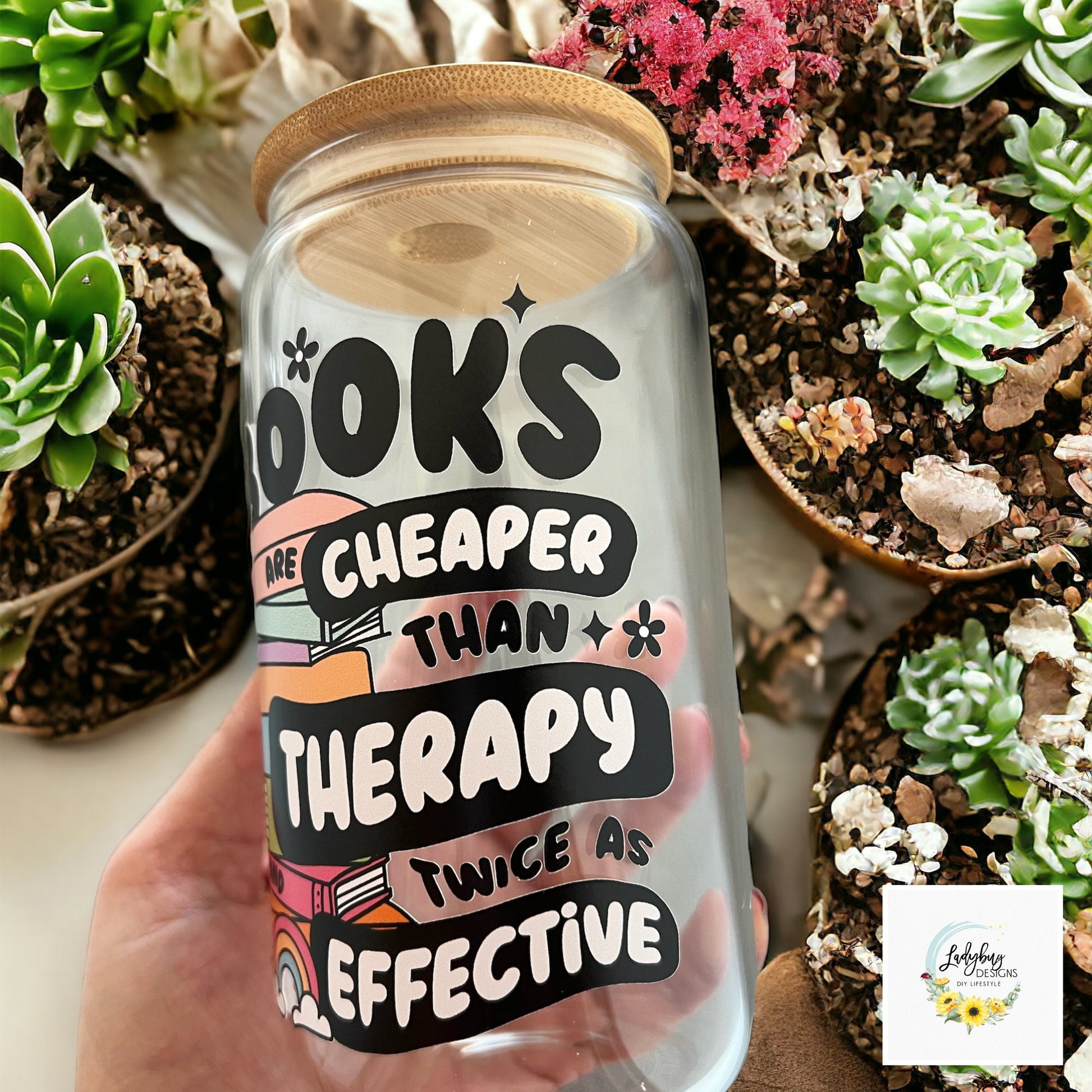 Book are Cheaper than Therapy Glass Can 16oz with Bamboo Lid and Plastic Straw. Ideal for iced coffee and perfect as a book lover&#39;s gift. Shown on a cozy reading table with a book and coffee cup, highlighting its stylish design.