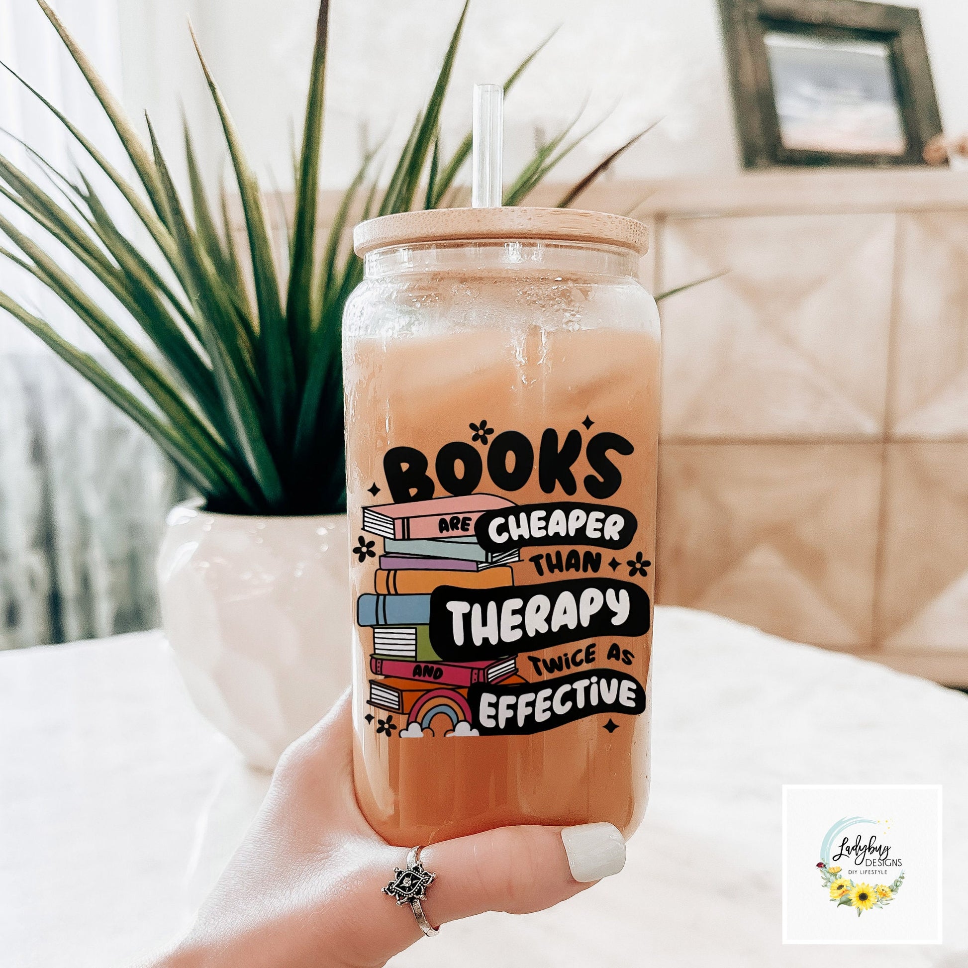 Book are Cheaper than Therapy Glass Can 16oz with Bamboo Lid and Plastic Straw. Ideal for iced coffee and perfect as a book lover&#39;s gift. Shown on a cozy reading table with a book and coffee cup, highlighting its stylish design.