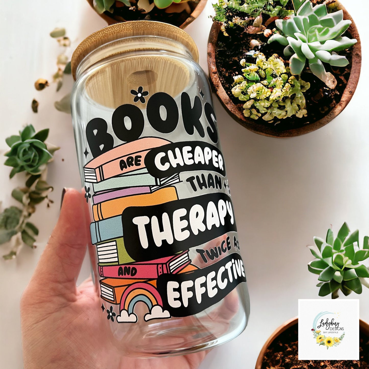 Book are Cheaper than Therapy Glass Can 16oz with Bamboo Lid and Plastic Straw. Ideal for iced coffee and perfect as a book lover&#39;s gift. Shown on a cozy reading table with a book and coffee cup, highlighting its stylish design.